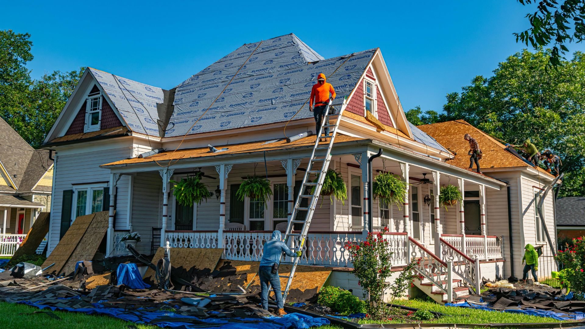 Workers replacing roof on white Victorian-style house with blue tarps