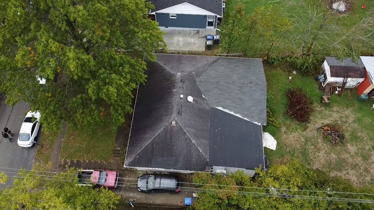 Aerial view of suburban house with dark roof, driveway, and surrounding trees